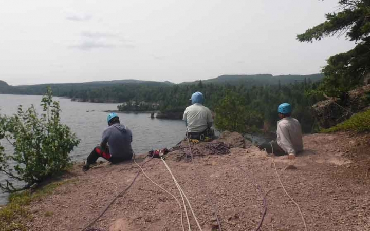 three people sit on a cliff above a body of water and prepare to rock climb on an outward bound expedition 